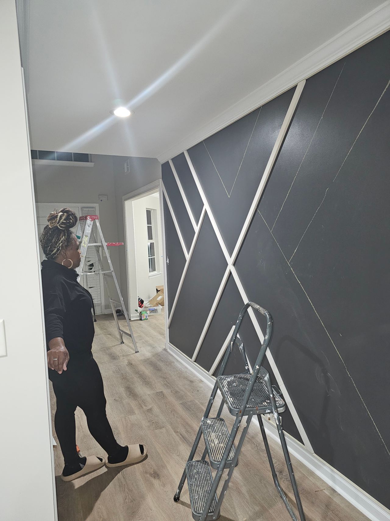 A hallway grey wall with some white battens fixed and the rest marked out; a person stands looking at it.