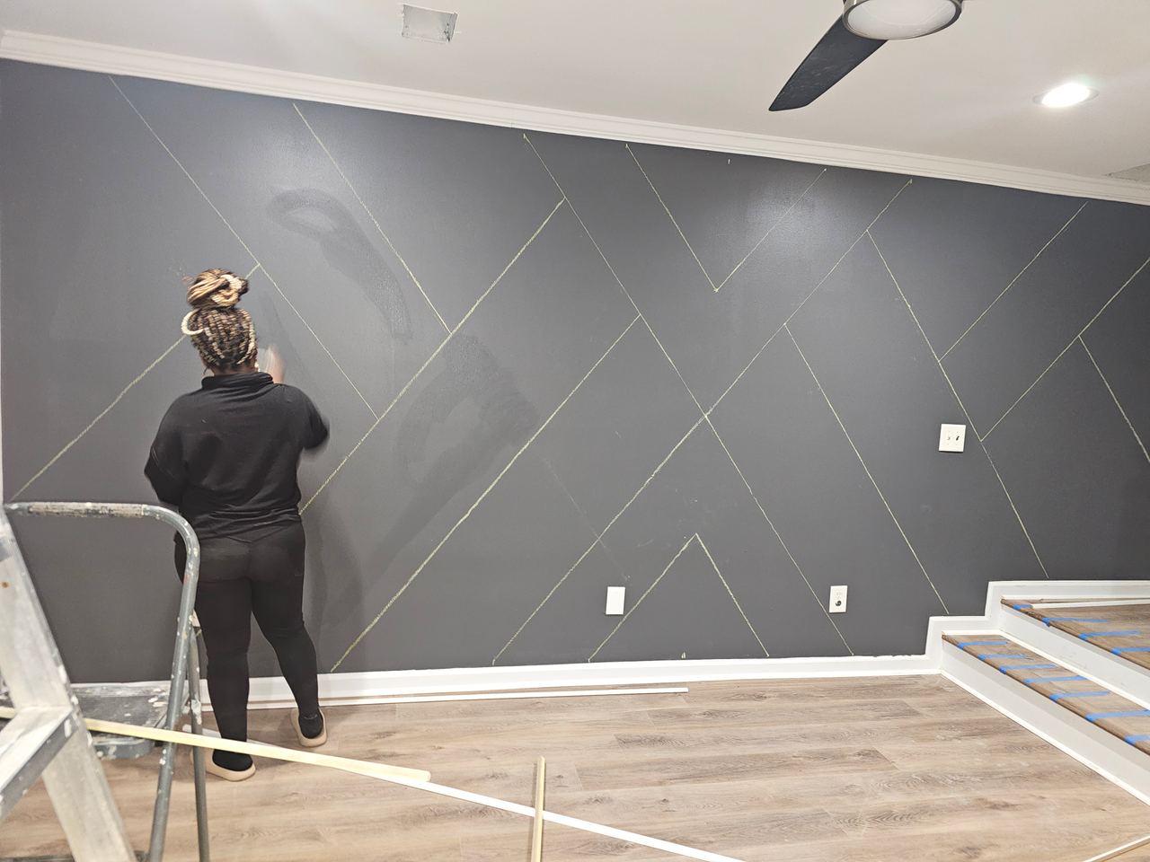 A dark grey wall with the batten pattern marked out in chalk lines; Mrs. Jones works at it working on the completion.